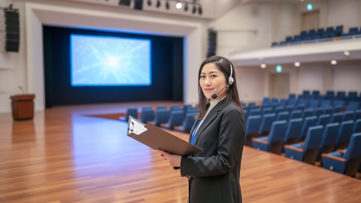 Female event staff preparing for a corporate event. Wearing a headset and holding documents, managing operations in a large hall.