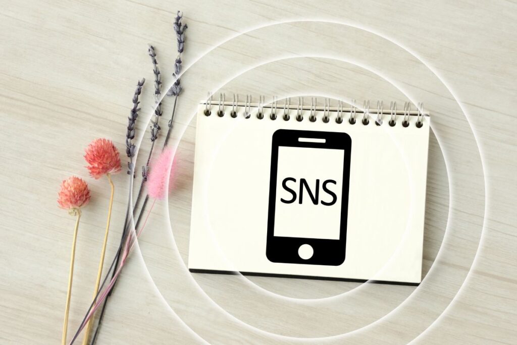 「SNS」とスマートフォンのイラストが描かれたノートと花が並ぶ写真。SNSを使った情報発信やコンテンツ配信のイメージ。