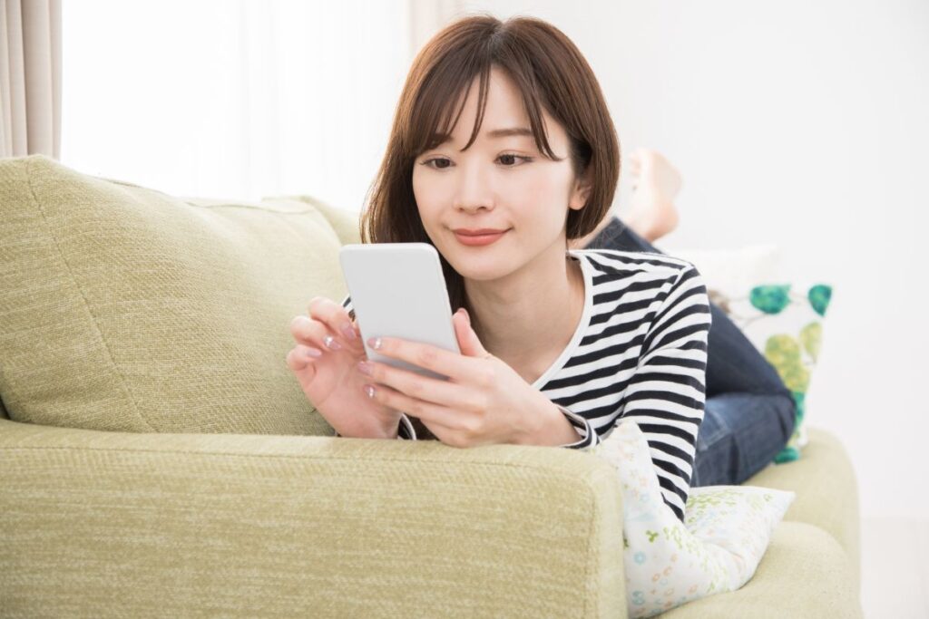 ソファに横になりスマホで検索し、情報収集やネットリサーチを行う女性のイメージ。