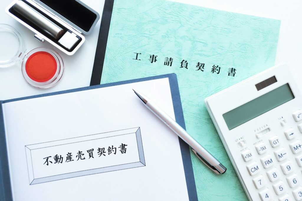 不動産売買契約書と工事請負契約書、印鑑、電卓、ペンが机の上に並べられている。不動産取引の契約手続きをイメージした写真。