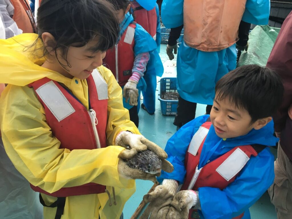 子どもたちが漁船の上で魚を手に取り、笑顔で触れ合っている漁師体験イベントの様子。地域の自然とふれあう楽しさを伝える写真。