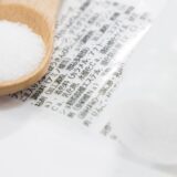 A photo showing white powdered additives on a wooden spoon over a Japanese food label, symbolizing awareness of food additives.