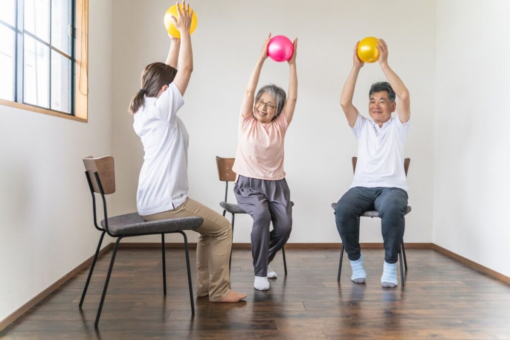 椅子に座ってボールを使いながらエクササイズをする高齢者たち。笑顔で楽しく体を動かしている様子。