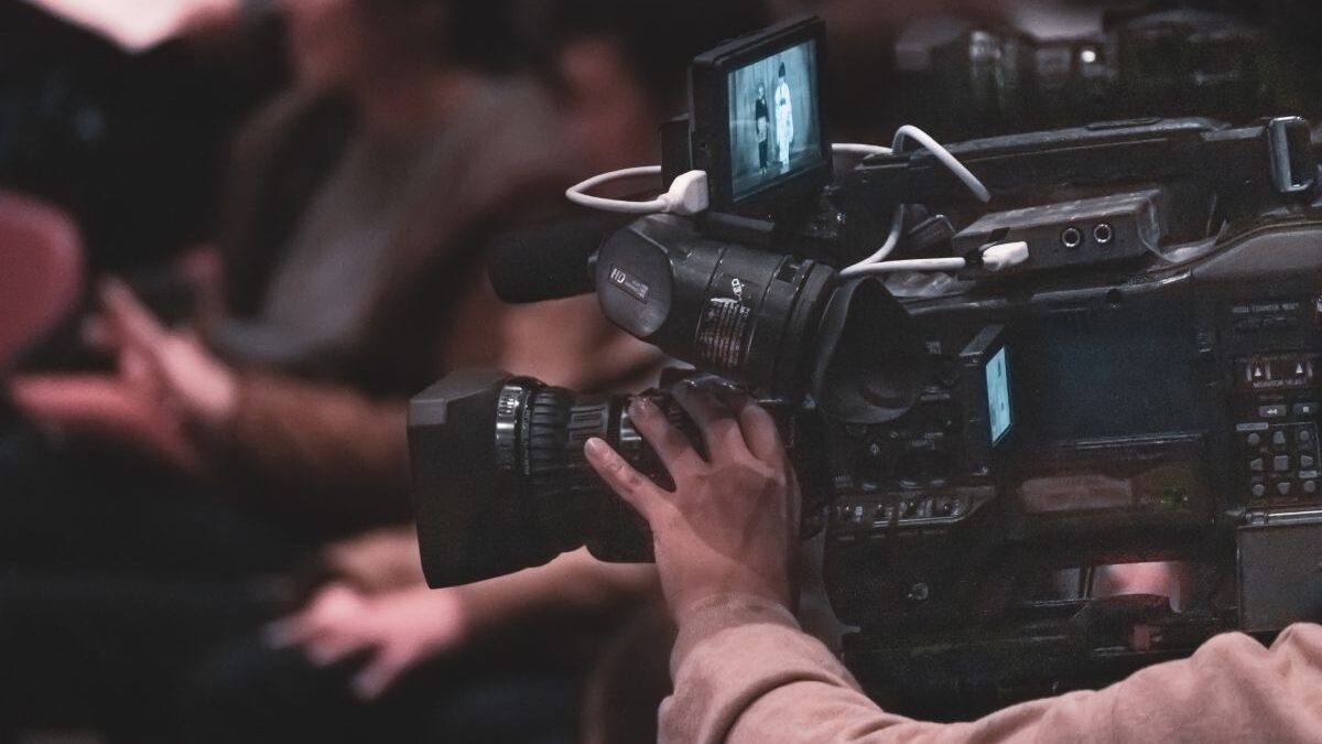 A professional videographer filming an event with a large video camera while the audience sits in the background.