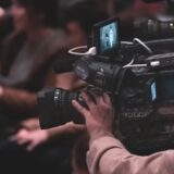 A professional videographer filming an event with a large video camera while the audience sits in the background.