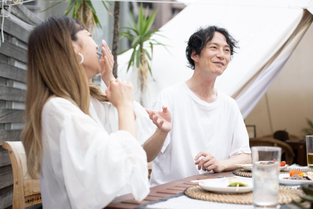屋外のテラスで食事をしながら笑い合うカップル。自然な笑顔と軽やかな雰囲気が伝わる。
