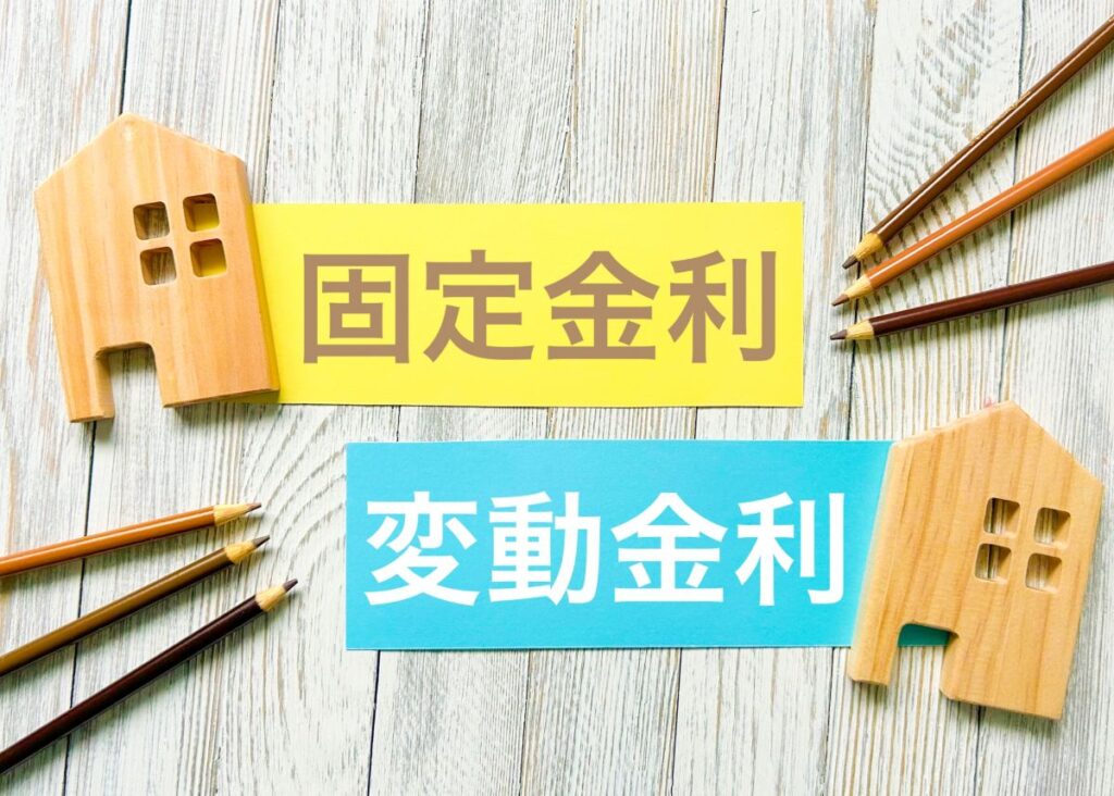木製の家の模型と鉛筆の横に「固定金利」「変動金利」と書かれたカードが並ぶ写真。金利タイプを比較するイメージ。