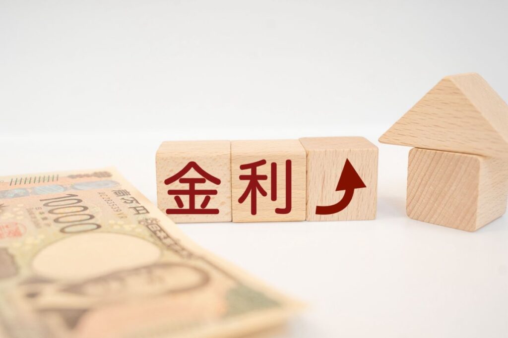 木製ブロックに「金利」と上向き矢印が描かれ、隣に一万円札と家の模型が置かれた写真。金利上昇をイメージしている。
