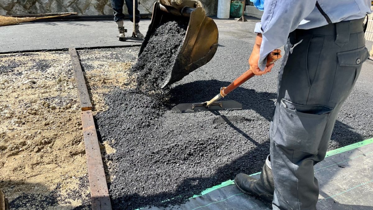 Workers paving a parking lot with asphalt using an excavator and shovels, showing the process of asphalt pavement construction.