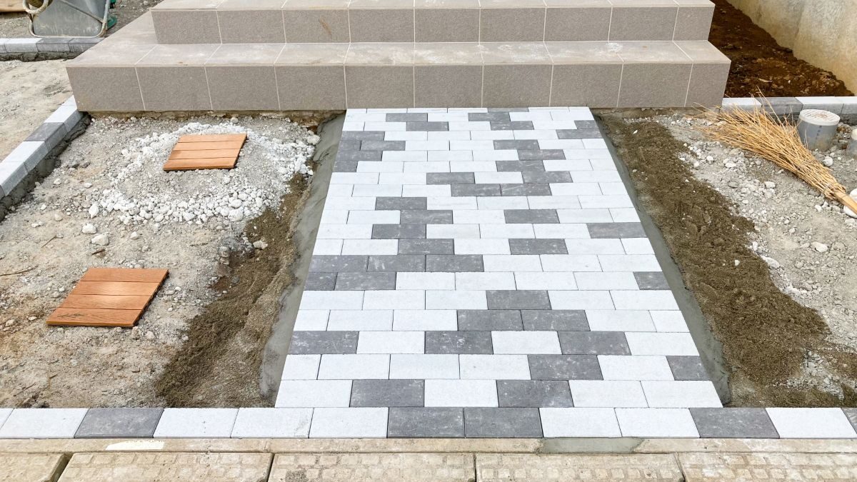 A partially completed exterior construction scene showing paving blocks being installed on the front approach.