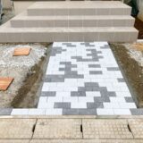 A partially completed exterior construction scene showing paving blocks being installed on the front approach.