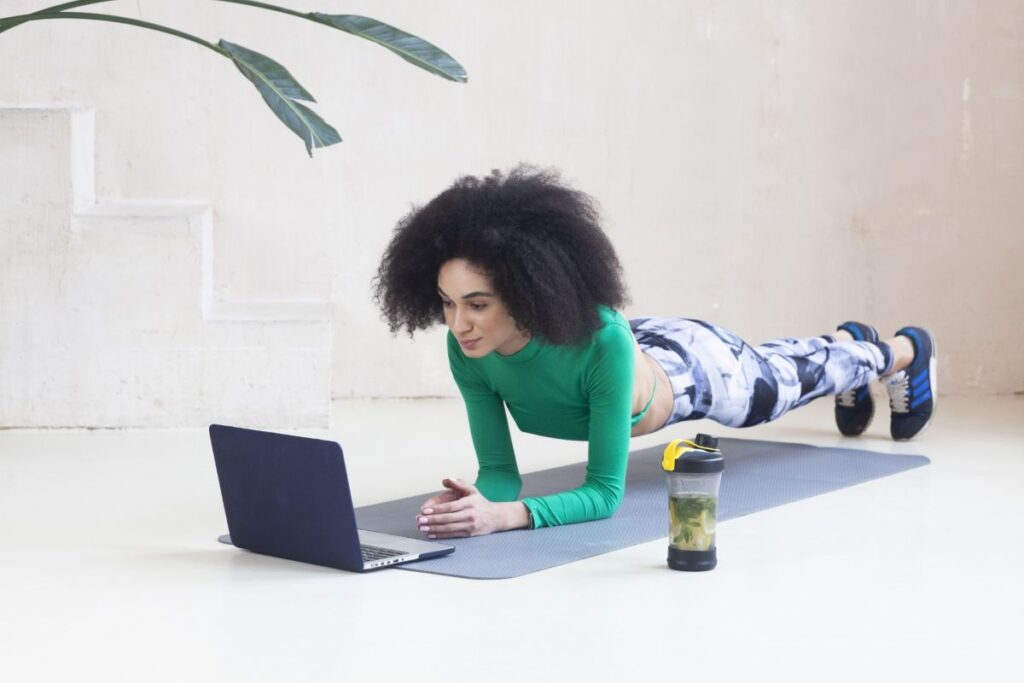 ヨガマットの上でプランクを行い、パソコン画面の指示を見ながらトレーニングしている女性の画像。