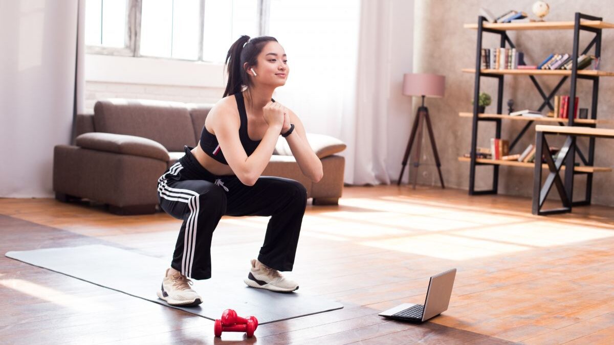 A woman doing squats at home while following an online workout video on a laptop.