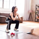 A woman doing squats at home while following an online workout video on a laptop.