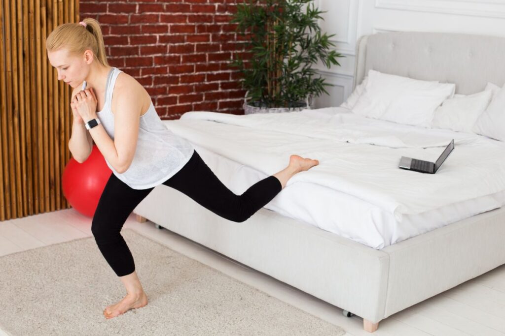 自宅のベッドを使って片脚エクササイズに取り組む女性が、オンラインレッスンを見ながらバランスを取っている画像。