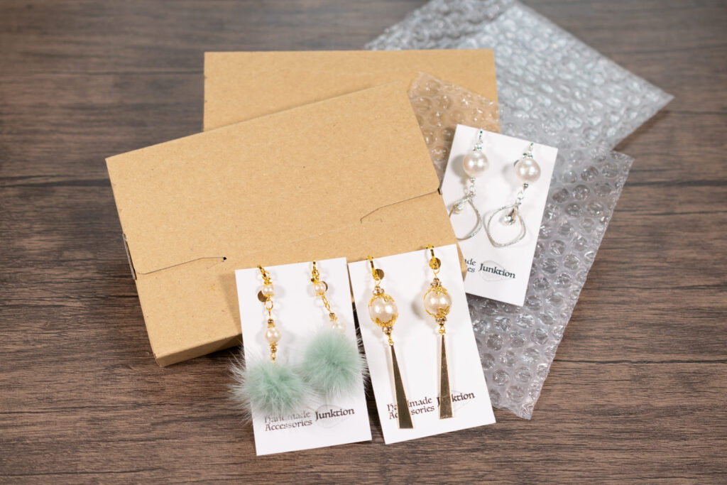 ネット販売の発送準備。段ボールの小箱、気泡緩衝材（プチプチ）、そして丁寧に台紙にセットされたハンドメイドのピアス3種類。
