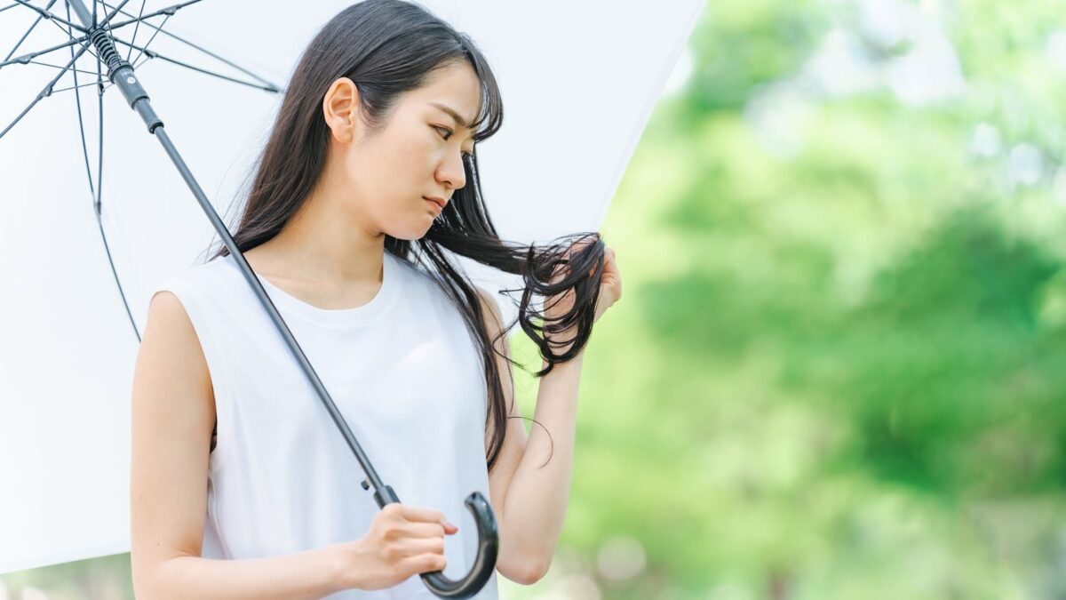 屋外で白い傘をさし、髪の毛先のパサつきやダメージを心配そうに見つめる若い女性。