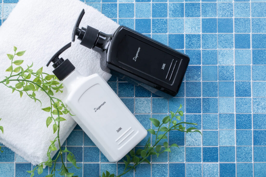 青いタイル張りの浴室に置かれた、シンプルでおしゃれな白と黒のシャンプー＆トリートメント用詰め替えディスペンサーボトルとタオル。