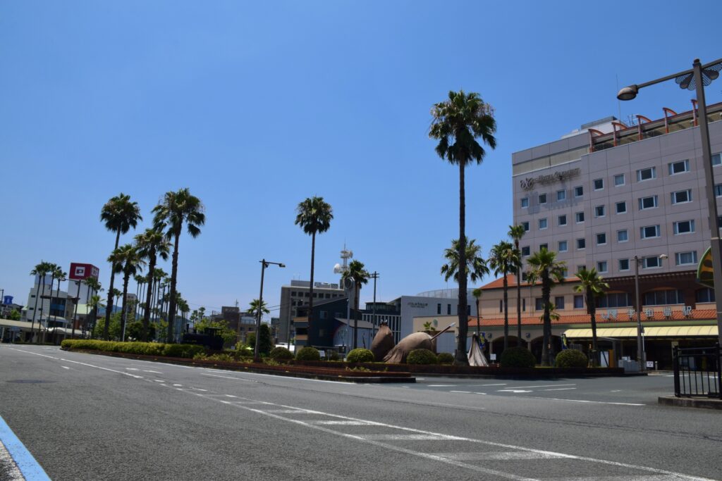 青空の下、南国風のヤシの木が立ち並ぶJR宇和島駅前のロータリーと駅舎の風景