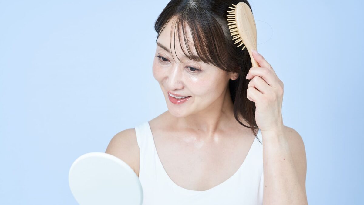 白いタンクトップを着た女性が、笑顔で手鏡を見ながら木製のヘアブラシで髪をとかしている様子。健康な髪と丁寧な頭皮ケアの習慣をイメージさせる写真。