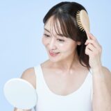 白いタンクトップを着た女性が、笑顔で手鏡を見ながら木製のヘアブラシで髪をとかしている様子。健康な髪と丁寧な頭皮ケアの習慣をイメージさせる写真。