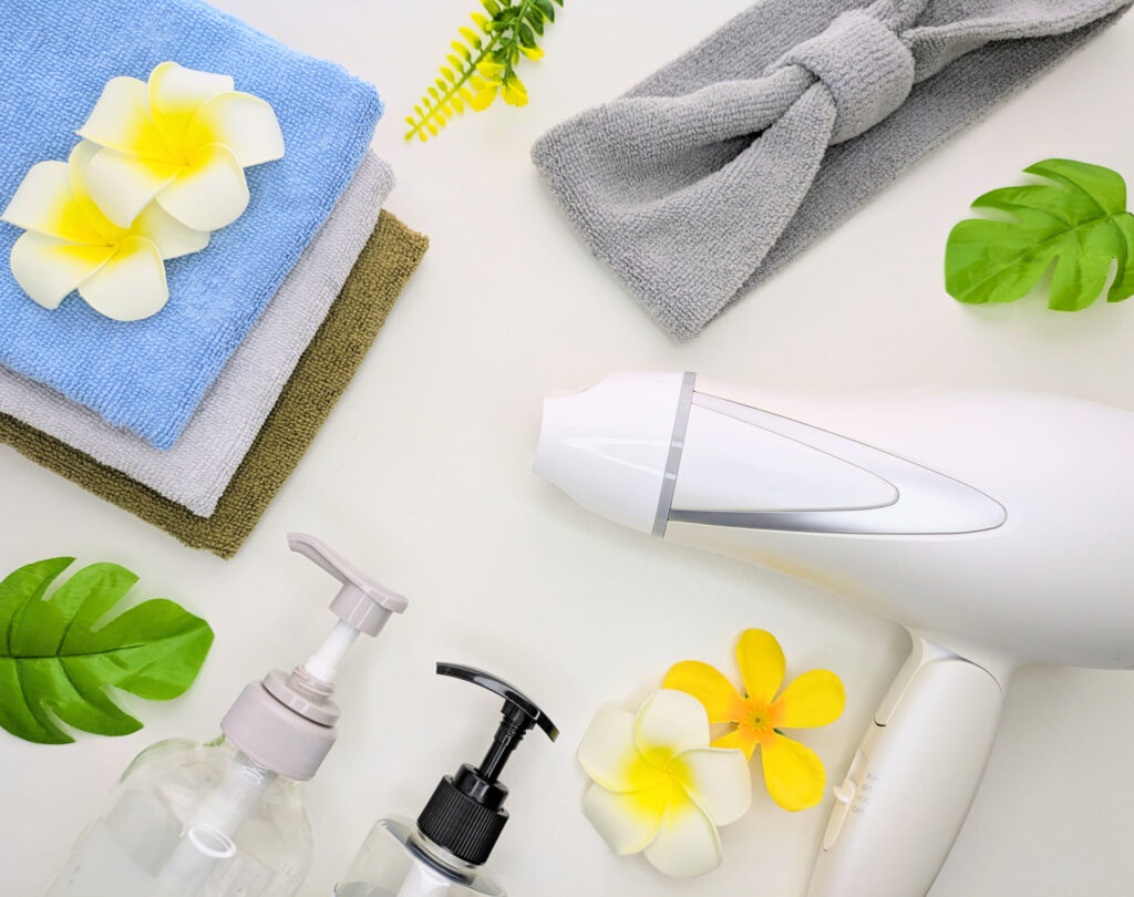 白いヘアドライヤー、積み重ねたタオル、ヘアバンド、透明なポンプボトル、プルメリアの花が白背景に並んだヘアケア用品のフラットレイ。