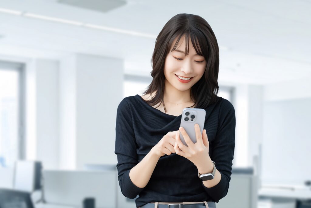 明るいオフィスでスマートフォンを操作し、笑顔を見せる若い女性。リスティング広告のターゲットユーザーや、検索意図を考える様子をイメージ。
