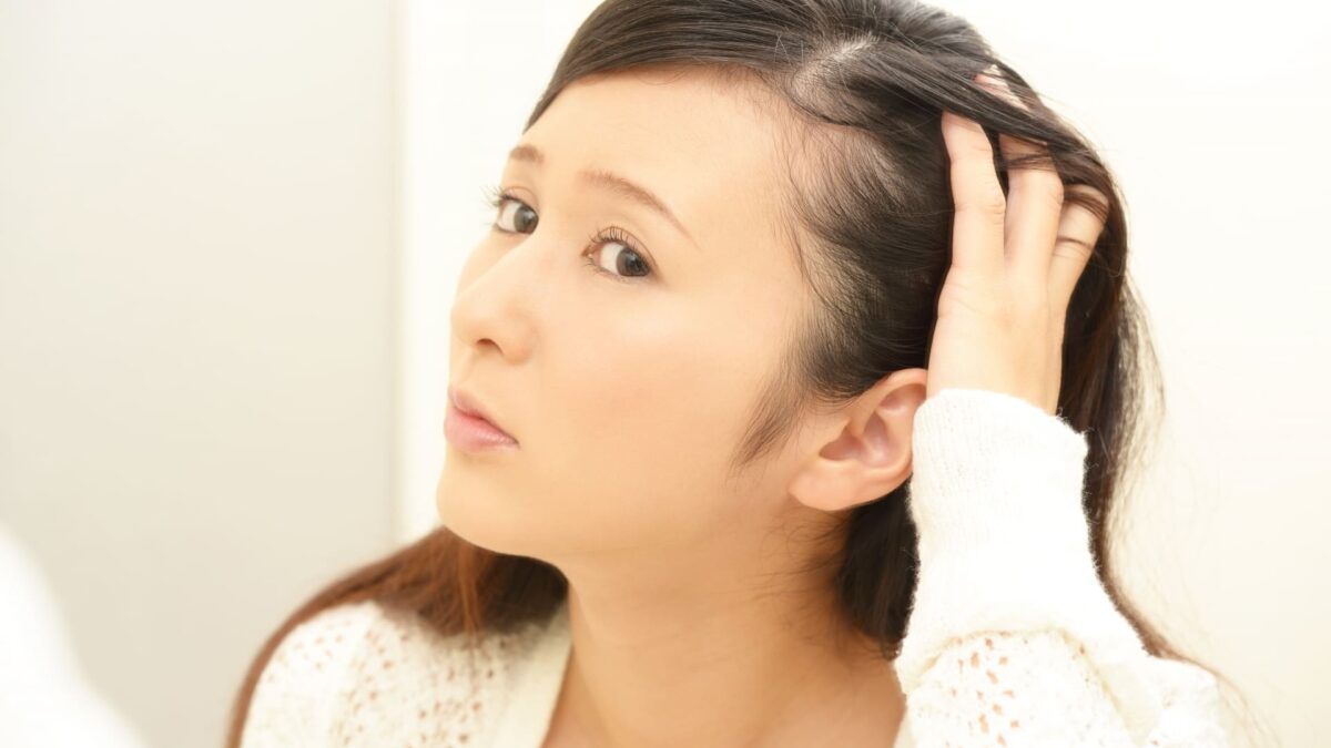 鏡を見ながら自分の髪や頭皮の状態を不安そうにチェックする日本人女性。薄毛、抜け毛、白髪などの髪の悩みを気にする様子