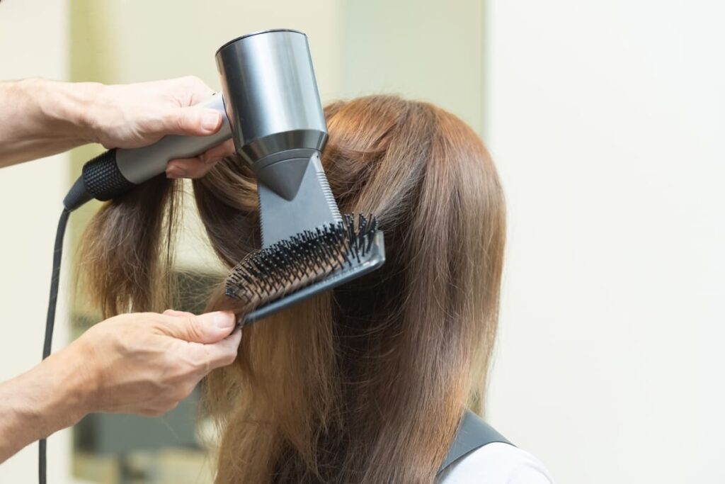 美容室で美容師がヘアドライヤーとブラシを使い、女性の髪を丁寧にブローして髪質改善を行う様子