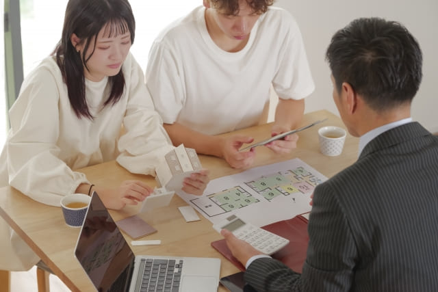 不動産会社の担当者とテーブルを囲み、家の模型と間取り図、電卓を見ながら真剣な表情で相談する30代の若い夫婦。