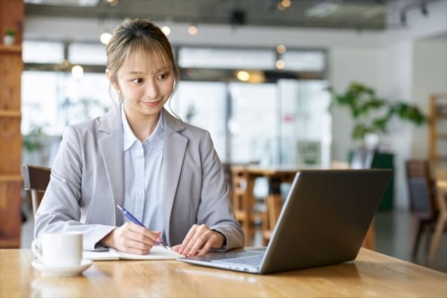 明るいカフェのようなオフィスで、ノートパソコンに向かいながら笑顔でノートにペンを走らせる、グレーのジャケットを着た若い女性ビジネスパーソン。自動化により生まれた時間でクリエイティブな戦略を練る様子