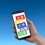 青空を背景に、手に持ったスマートフォンの画面上に赤・黄・青などのカラフルな「広告」という文字が並んでいる様子。モバイル環境での広告露出のイメージ。