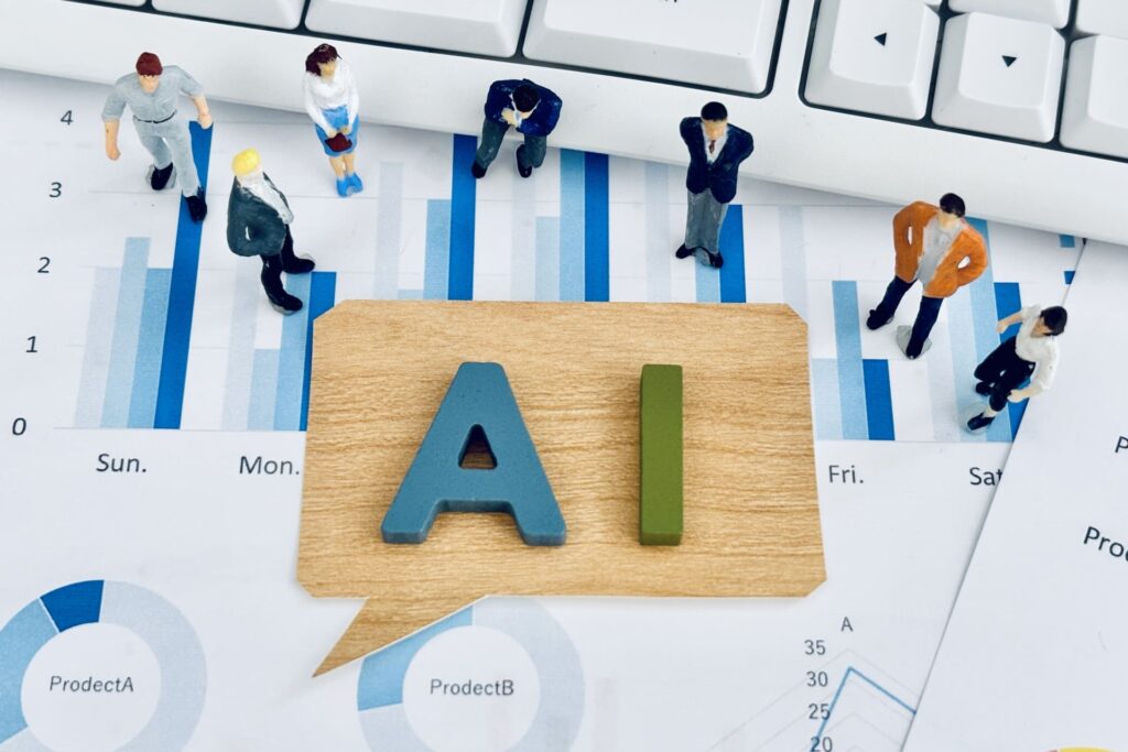 棒グラフや円グラフなどのデータレポートの上に置かれた、「AI」の文字が書かれた木の吹き出し型ブロックと、周囲に立つビジネスパーソンのミニチュアフィギュア。広告運用のデータ分析とAIによる自動化のメタファー