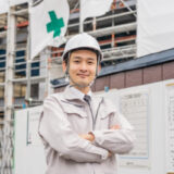 A young male site manager (foreman) in a hard hat smiling with crossed arms, with a construction site in the background.