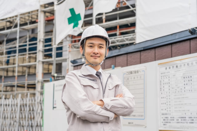 A young male site manager (foreman) in a hard hat smiling with crossed arms, with a construction site in the background.