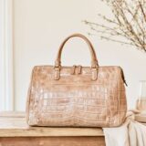A tan crocodile leather bag placed on a wooden table near a sunlit window, styled with branches and a glass of wine.