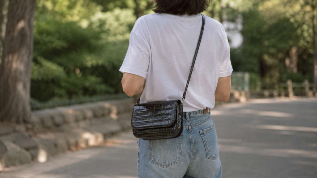 白Tシャツとデニムを合わせた人物が、黒いクロコダイル革のショルダーバッグを提げて屋外を歩く後ろ姿