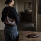 A person in denim and a plain black top carrying a crocodile leather shoulder bag in a dark-toned interior, with a matching crocodile leather wallet resting on a wooden table nearby